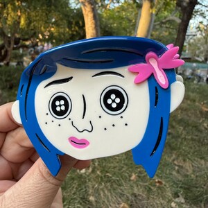 May include: A blue and white cartoon character brooch with button eyes and a pink flower. The character has blue hair and freckles. The brooch is held up against a background of green trees and sunlight.