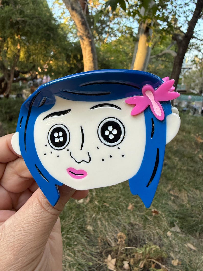 May include: A blue and white cartoon character brooch with button eyes and a pink flower. The character has blue hair and freckles. The brooch is held up against a background of green trees and sunlight.