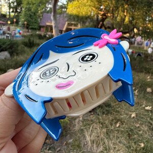 May include: A blue and white hair claw clip featuring a cartoon face with blue hair, black eyes, and a pink flower accent. The clip has a claw design for securing hair and is made of plastic.
