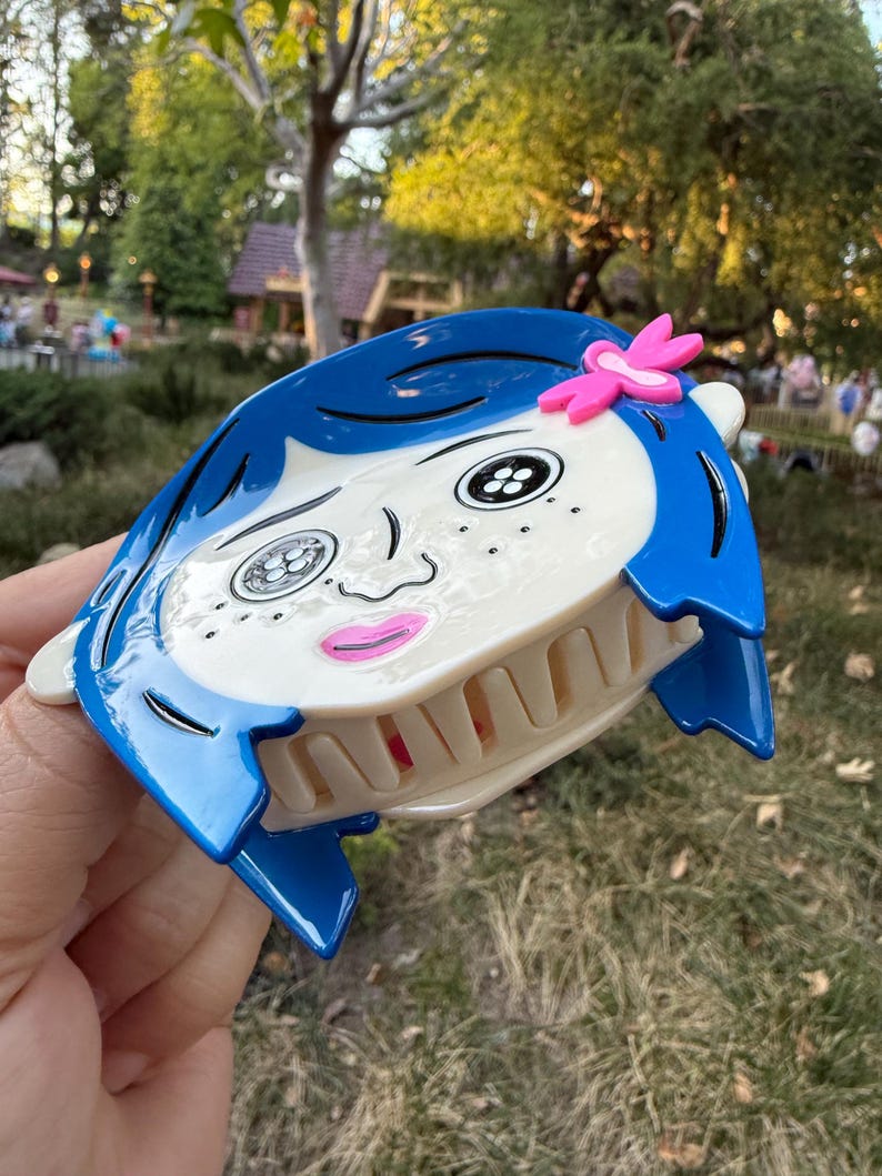 May include: A blue and white hair claw clip featuring a cartoon face with blue hair, black eyes, and a pink flower accent. The clip has a claw design for securing hair and is made of plastic.