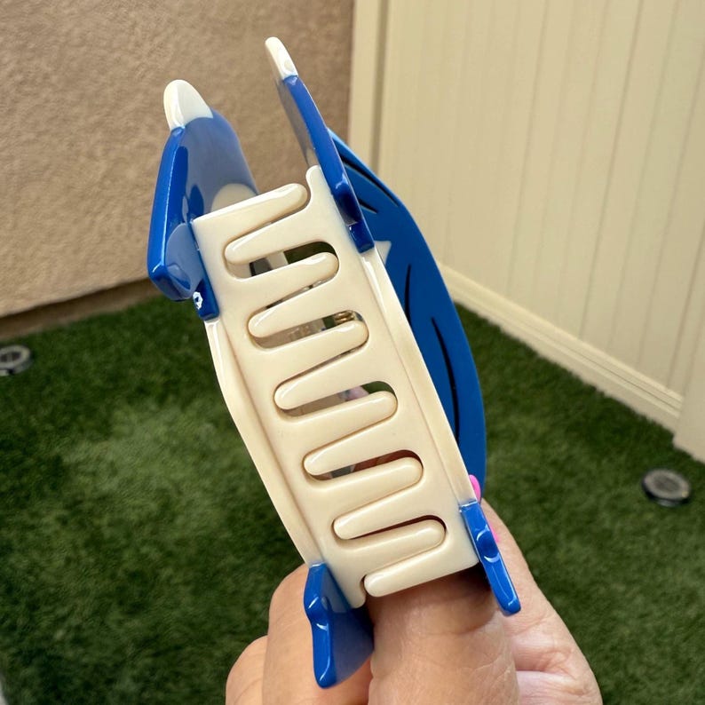 May include: A blue and white hair claw clip. The clip has a curved design with a series of interlocking teeth in a cream color. The sides of the clip are blue with white accents. The clip is held by a hand.