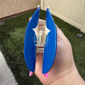 May include: A blue hair claw clip with a white accent and pink tips. The clip has a spring mechanism and is held open. This hair accessory is designed to hold hair in place.