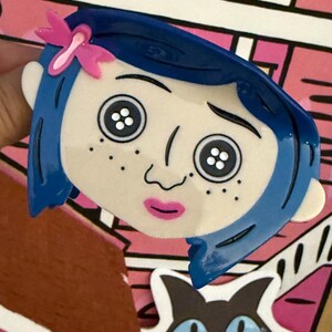 May include: A blue-haired cartoon character face with button eyes and a pink bow. The character has freckles and a pink mouth. The background features a pink and white house design and a black cat.