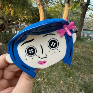 May include: A blue and white character face with black button eyes, freckles, and a pink bow. The character has blue hair and a pink mouth. The design is a stylized representation of a character from a movie.