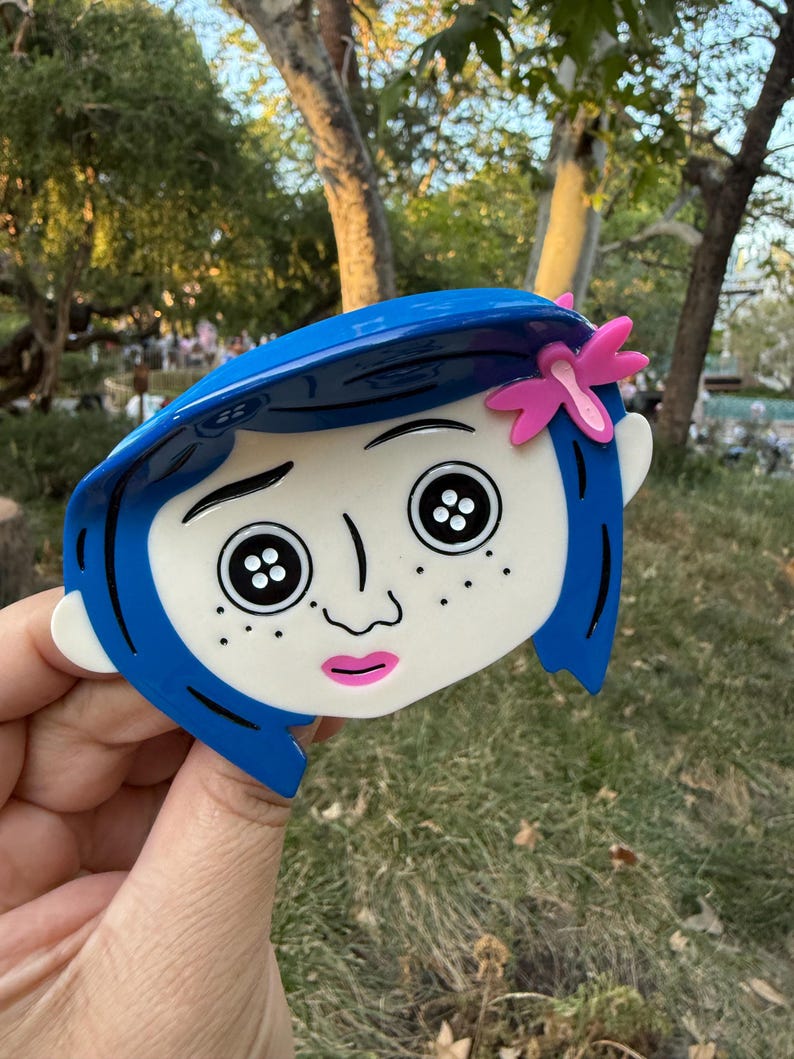 May include: A blue and white character face with black button eyes, freckles, and a pink bow. The character has blue hair and a pink mouth. The design is a stylized representation of a character from a movie.
