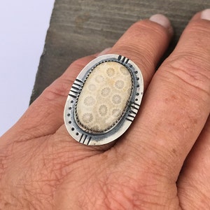 Pode incluir: Um anel de prata com uma pedra de coral fossilizado branco engastada em forma oval. O anel tem uma borda decorativa com acentos pretos.