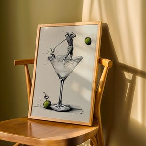 May include: A framed art print featuring a golfer swinging a club inside a martini glass. The artwork is in black and white with green accents, including a golf ball and a garnish. The print is displayed on a wooden chair.