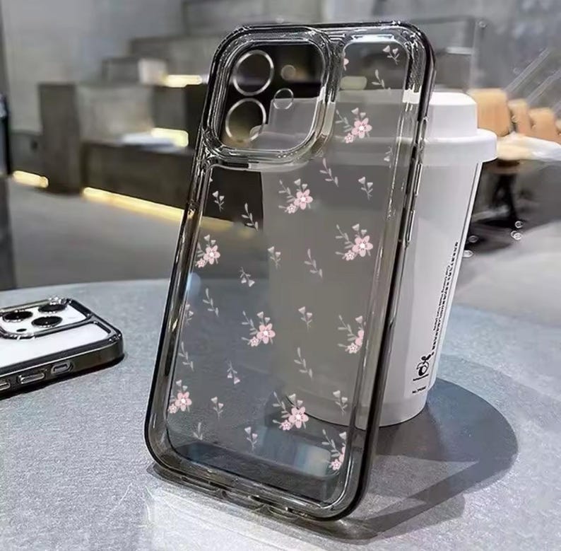 Op de afbeelding: Een transparante telefoonhoes met een bloemenpatroon. De hoes heeft een verhoogde rand rond de camera lens en een transparante achterkant.