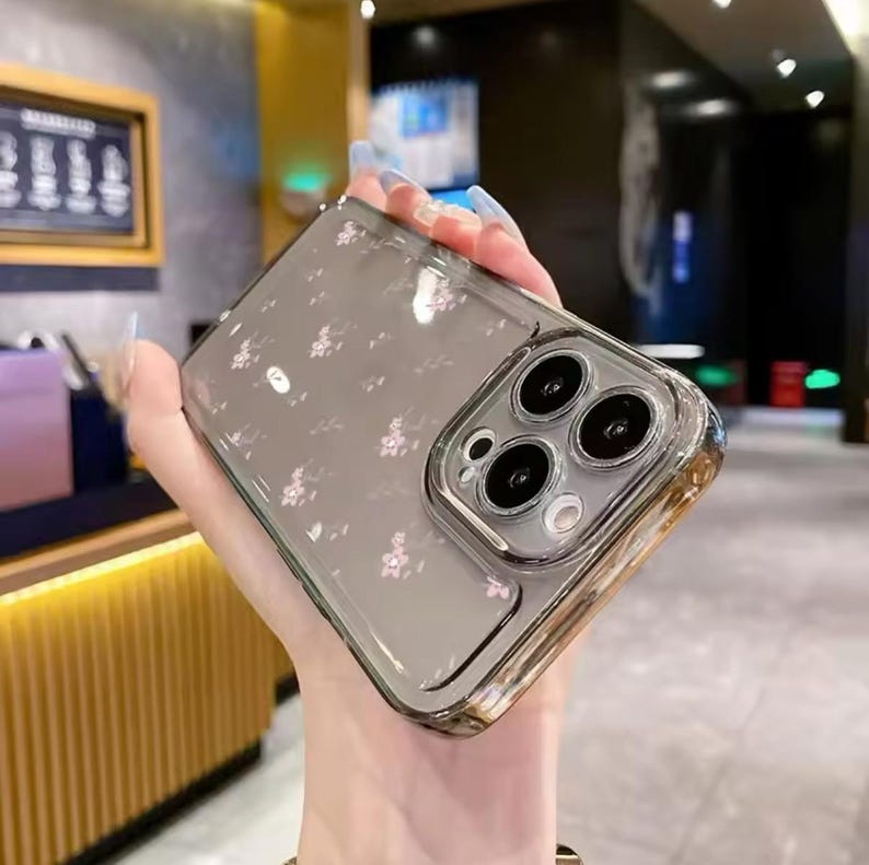 Op de afbeelding: Een transparante telefoonhoes met een bloemenpatroon en een zwarte camera lens cover. De hoes is ontworpen voor een iPhone 13 Pro.