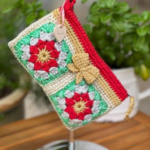 May include: A handmade crochet clutch featuring a vibrant floral pattern in red, green, and gray. The clutch has a red border and a gold-colored bow closure. A small wooden heart with text hangs from the top.