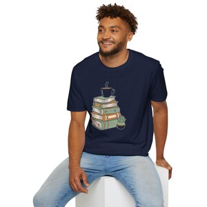May include: Navy blue t-shirt featuring a graphic of a stack of books, a coffee cup, and a small potted plant. The design is in shades of green, orange, and brown. The shirt is made of a soft material.