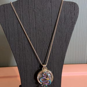 Puede incluir: Un collar de cadena de plata con un colgante redondo que presenta una serpiente iridiscente enroscada.