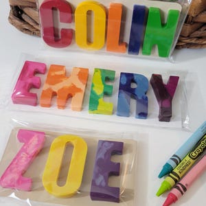 May include: Colorful crayon letters spelling out the names "Colin", "Emery", and "Zoe". Each name is packaged separately. Crayola crayons are visible in the background. The letters are in various colors, including red, orange, yellow, green, blue, and purple.