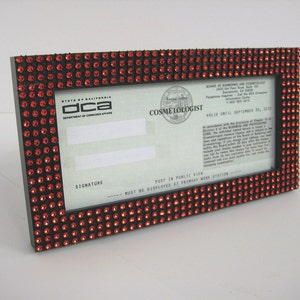 May include: A black frame with a red rhinestone border surrounds a California Cosmetologist license. The license is valid until September 30, 2015. The text "dca" is visible in the upper left corner of the license.