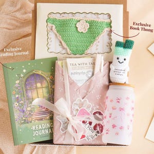Puede incluir: Una caja de regalo con un diario de lectura verde, un tanga de ganchillo verde, un tarro blanco con diseños florales rosas y una muñeca de puerro de ganchillo. El diario tiene un diseño de ventana floral. La caja está envuelta en papel rosa con una cinta blanca.
