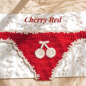 May include: A red crochet bikini bottom featuring a white crocheted cherry motif in the center and a cream-colored scalloped trim along the edges. The text Cherry Red is printed in red above the garment on a white background with floral patterns.