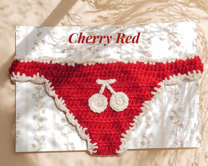 May include: A red crochet bikini bottom featuring a white crocheted cherry motif in the center and a cream-colored scalloped trim along the edges. The text Cherry Red is printed in red above the garment on a white background with floral patterns.