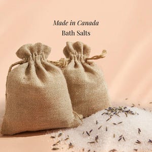 May include: Two burlap drawstring bags of bath salts with the text "Made in Canada" and "Bath Salts". A pile of white bath salts and lavender buds are in front of the bags. The background is a light peach color.
