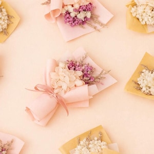 May include: Overhead view of several small bouquets of dried flowers. Each is wrapped in pink or yellow paper and tied with ribbon. The flowers are a mix of white, purple, and beige, creating a delicate, natural aesthetic.
