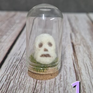 以下が含まれることがあります： 暗い茶色の顔の特徴を持つ小さな白いフェルトスカルが、透明なガラスの鐘の中に収められています。 スカルは、コルクベースの上に、緑の苔のベッドの上にあります。 鐘は木製の表面にあります。