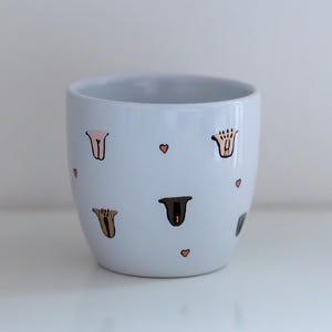 Może przedstawiać: Jasnoniebieska ceramiczna doniczka o zaokrąglonym kształcie. Ozdobiona abstrakcyjnymi kształtami w odcieniach brązu i różu oraz małymi różowymi sercami. Nadaje się do roślin domowych. Wymiary w centymetrach.