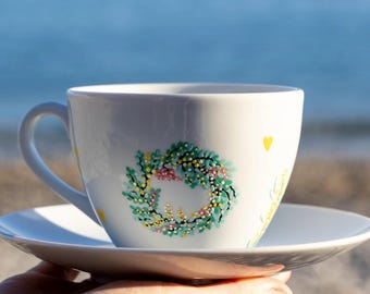 May include: White teacup and saucer set with a hand-painted wreath design. The wreath features green leaves, pink and yellow flowers, and small yellow dots. The cup is held in a hand against a blurred blue background.