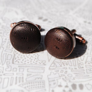 May include: A pair of brown leather cufflinks with a copper finish. The cufflinks have a textured surface and a circular shape.