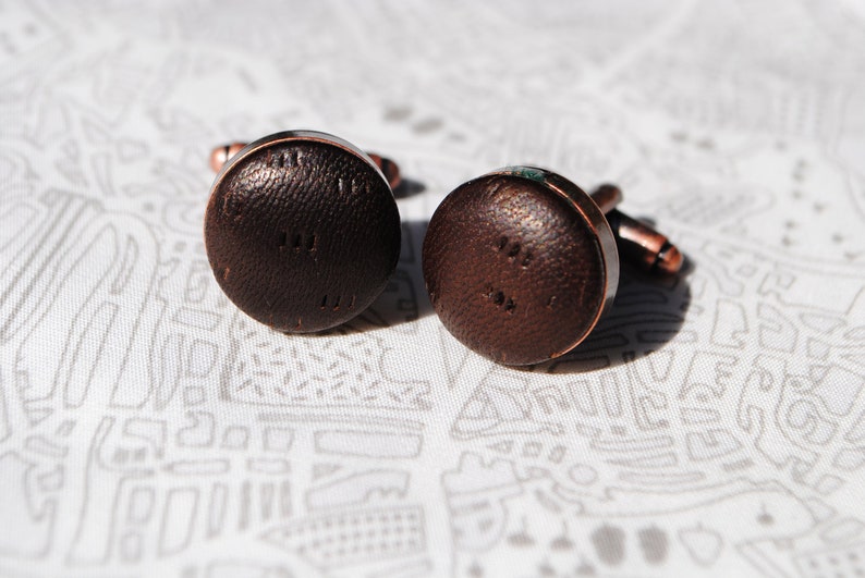 May include: A pair of brown leather cufflinks with a copper finish. The cufflinks have a textured surface and a circular shape.