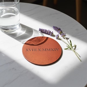 May include: A round, reddish-brown coaster with the Roman numeral date XVIII.X.MMXIV printed on it. A sprig of lavender and a glass of water are also visible. The coaster is on a white marble surface.