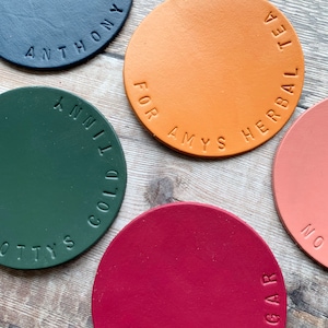 May include: Five round leather coasters in different colours and with text. The coasters are green, red, orange, black and pink. The text on the coasters reads "OTTY'S COLD", "NO SUGAR", "FOR AMYS HERBAL TEA", "ANTHONY", and "NO".