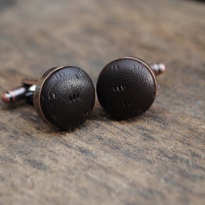 May include: A pair of cufflinks with a brown leather design. The cufflinks have a copper-colored metal base.