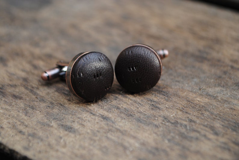 May include: A pair of cufflinks with a brown leather design. The cufflinks have a copper-colored metal base.