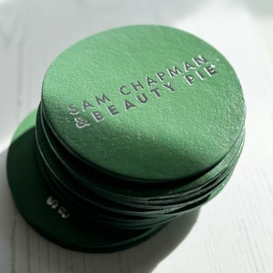 May include: A stack of green round coasters with the text "Sam Chapman & Beauty Pie" in silver lettering.