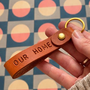May include: A tan leather keychain strap featuring the embossed text OUR HOME, secured with a gold-toned metal rivet and a matching circular gold-toned metal split ring for attaching keys, showcasing a smooth finish and visible leather texture.