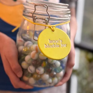 May include: A glass jar filled with colorful marbles, with a yellow tag that reads "Isaac's Marble Jar".
