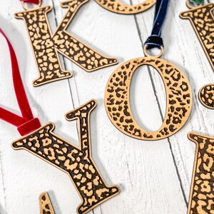 May include: Wooden letters in the shape of K, O, Y, and I, with a leopard print design. Each letter has a string attached to it, making them suitable for hanging as ornaments.