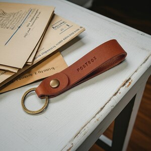 May include: A tan leather keychain with the word "POSTBOX" embossed on the strap. The keychain has a brass-coloured ring and a button closure. The keychain is on a white surface with vintage envelopes in the background.