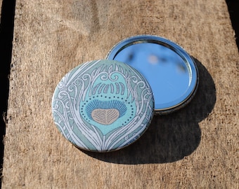 May include: A compact mirror with a peacock feather design. The mirror has a silver trim and a blue and green patterned design.