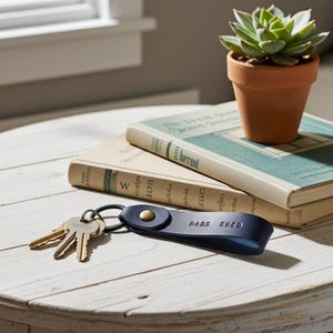 May include: A navy blue leather keychain with the words "DADS SHED" embossed on it, next to a set of keys. The keychain is on a white wooden table, with two vintage books and a small potted succulent in the background.