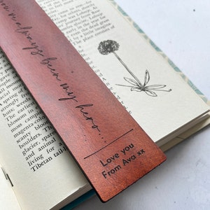 May include: A brown leather bookmark with the inscription "we've always been my hero..." in elegant script. The bottom of the bookmark reads "Love you From Ava xx". The bookmark is placed in an open book.