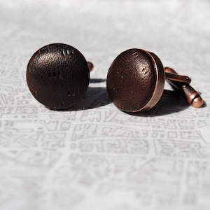 May include: A pair of brown cufflinks with a textured surface. The cufflinks are made of metal and have a copper finish.