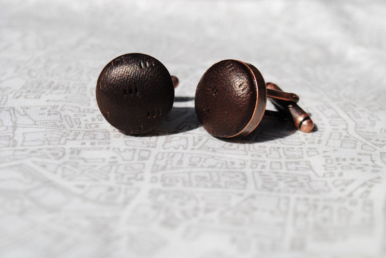 May include: A pair of brown cufflinks with a textured surface. The cufflinks are made of metal and have a copper finish.