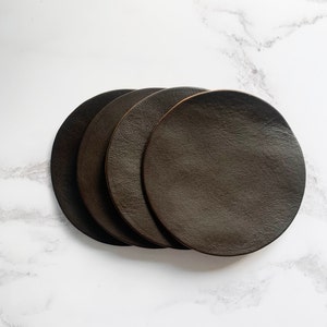 May include: Four brown leather coasters stacked on top of each other. The coasters are round and have a smooth, textured surface.