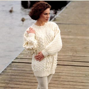 Puede incluir: Un suéter de punto crema de gran tamaño con intrincados detalles de trenzas y bolitas. El suéter se combina con leggings de color claro y botas marrones con cordones. Adecuado para climas fríos y ropa informal.