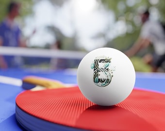 May include: A white ping pong ball with a decorative design featuring the number 8 in black, teal, and white. The ball is on a red ping pong paddle, with a blue table and blurred background of people playing.