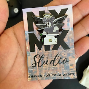 May include: A pin featuring a gray angel statue with black wings and a gold halo. The pin is on a card with the text "NX MX Studio" and "THANKS FOR YOUR ORDER" against a cityscape backdrop.