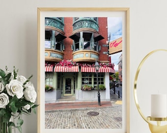 May include: A framed print of a building with a red brick facade, green trim, and a red and white striped awning. The building has ornate balconies and is adorned with flowers. A street lamp stands in front of the building on a cobblestone street.