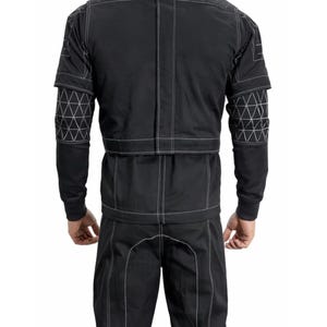 Op de afbeelding: Zwart jumpsuit met witte stiksels. De outfit heeft geometrische patronen op de mouwen en een gestructureerd ontwerp. De achteraanzicht toont het volledige kledingstuk, waarbij de getailleerde pasvorm en contrasterende stiksels worden benadrukt.