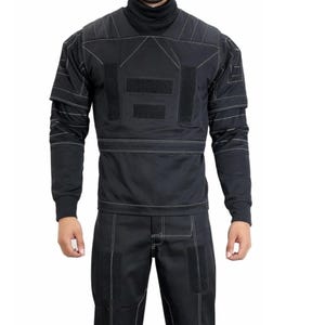 Op de afbeelding: Zwart tactisch uniform met een coltrui, lange mouwen en een bijpassende broek. Het uniform heeft witte stiksels en meerdere klittenband patches. Een functioneel en stijlvol ontwerp.