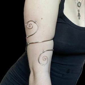 May include: Silver arm cuff with a spiral design. The cuff wraps around the upper arm and forearm, with a delicate, minimalist aesthetic. The jewellery is made of thin, flexible metal.